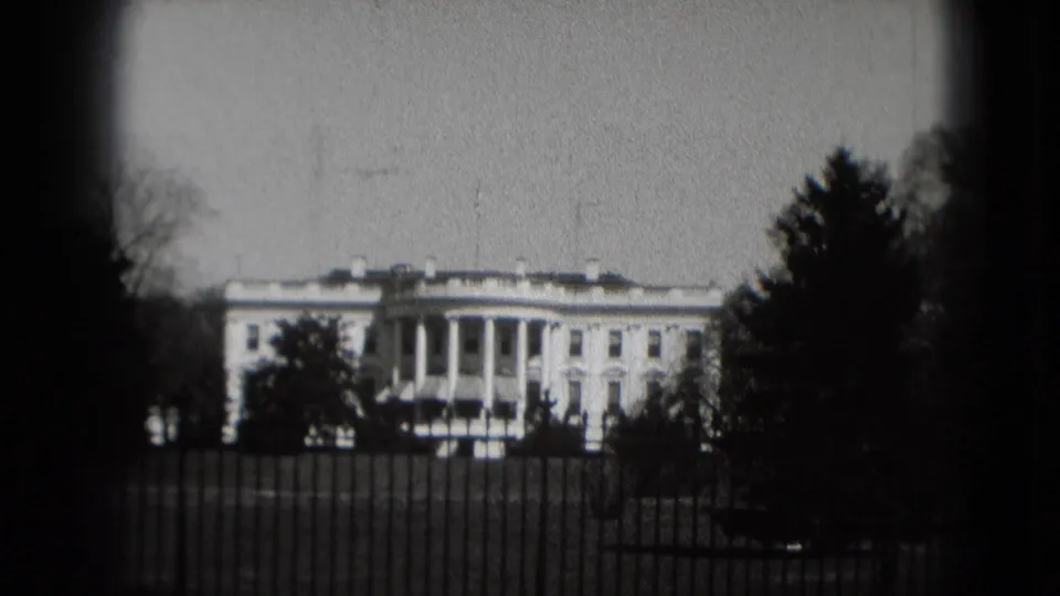 Black-and-white 8mm frame of the White House exterior in Washington, DC, with iron fence, lawn, trees, and soft focus.