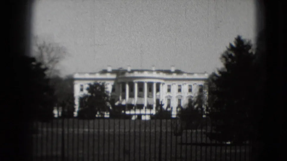 Grainy black-and-white 8mm view of the White House behind the fence, framed by trees and dark lens edges in Washington, DC.