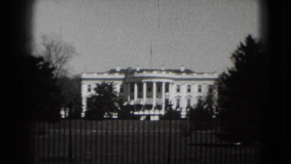 1975 8mm view of the White House in Washington, DC, framed by trees behind an iron fence on a cloudy day.