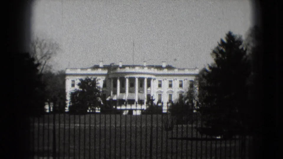 Black-and-white 8mm frame showing the White House in Washington, DC, beyond a foreground fence and winter-looking trees.
