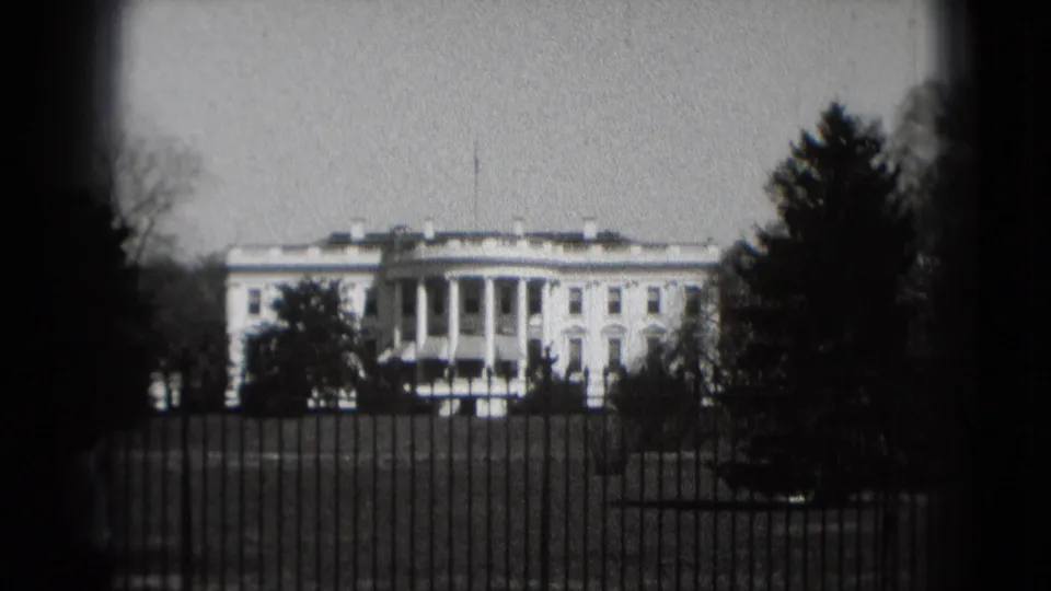 1975 home-movie frame of the White House behind the iron fence, bordered by trees and small-gauge film edges.