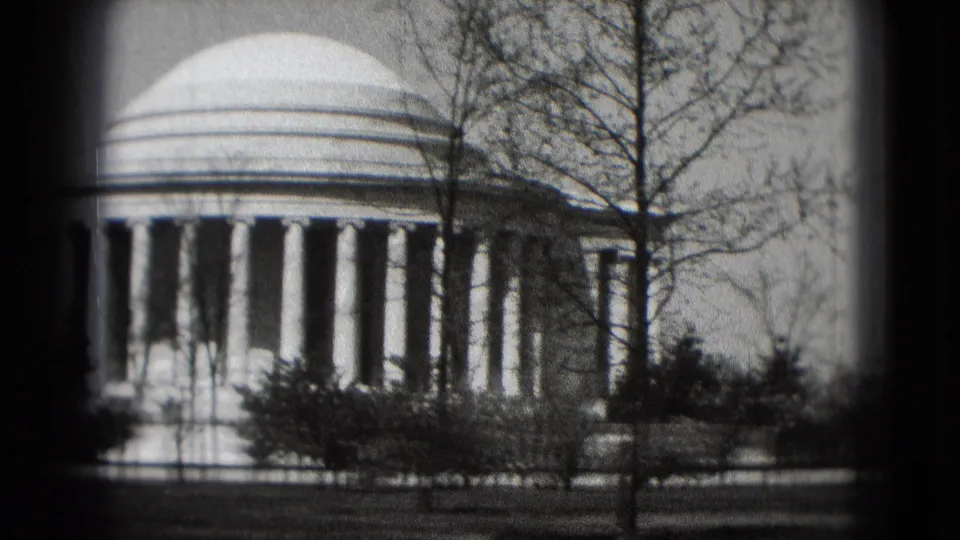 1975 home-movie frame showing the Jefferson Memorial columns and dome edge partly obscured by trees and foreground shrubs.