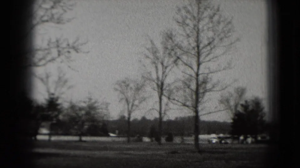 Black-and-white 1975 Washington, DC home-movie frame showing bare trees, open grass, distant cars, and dark film gate edges.