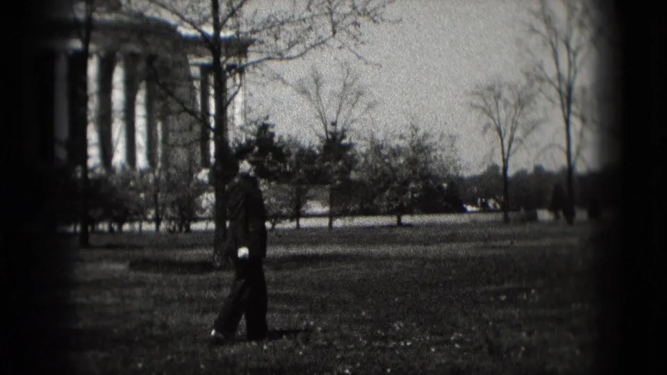 A distant adult figure walks across a grassy Washington, DC park, with classical columns, shrubs, bare trees, and cars in the background.