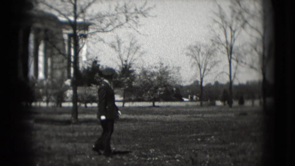 In 1975 Washington DC, a uniformed man walks across a grassy area with the Lincoln Memorial partly visible behind bare trees.