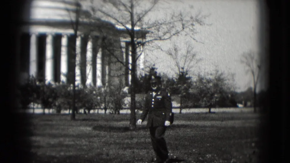 A uniformed man walks across the lawn in front of the Lincoln Memorial in Washington DC, seen through grainy 8mm footage.