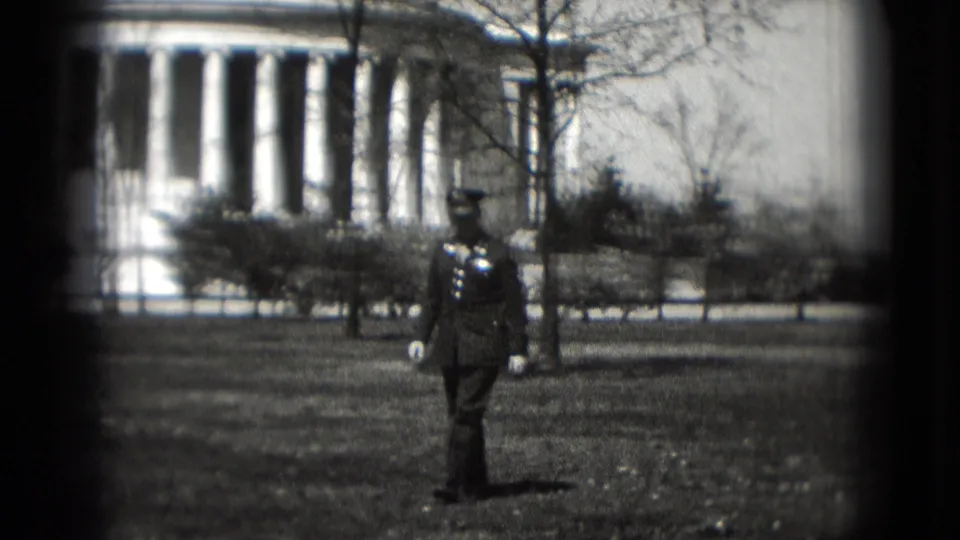 A closer view shows the uniformed man on the Washington DC memorial lawn, with Lincoln Memorial columns and shrubs behind him.