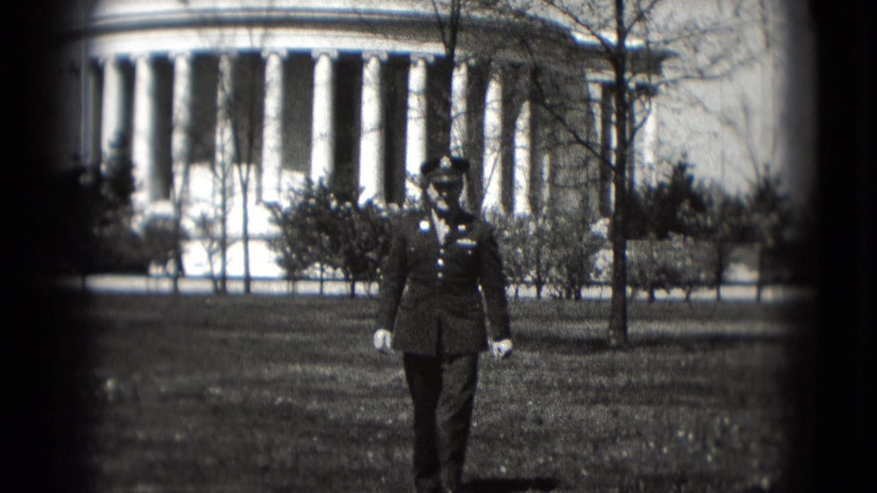 A uniformed serviceman walks toward the camera on a grassy lawn in Washington DC, with a neoclassical columned building in the background.