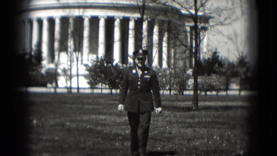 A uniformed serviceman walks toward the camera on a grassy lawn in Washington DC, with a neoclassical columned building in the background.