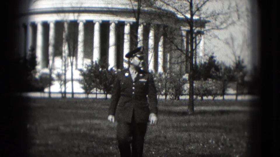 The serviceman remains centered while walking across the grass, with bare trees and a large columned monument visible behind him.