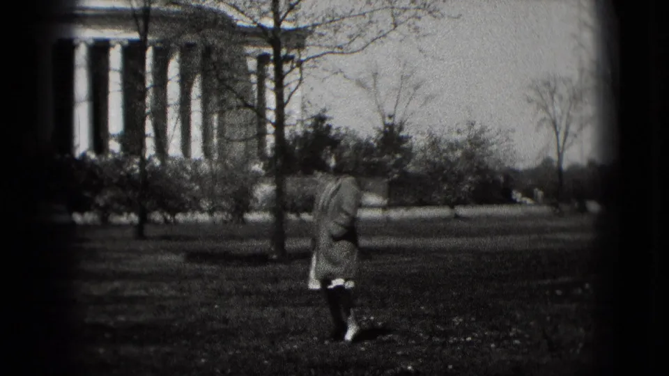 In Washington, D.C., a woman in a long coat walks across a grassy lawn near the Jefferson Memorial in black-and-white 8mm footage.