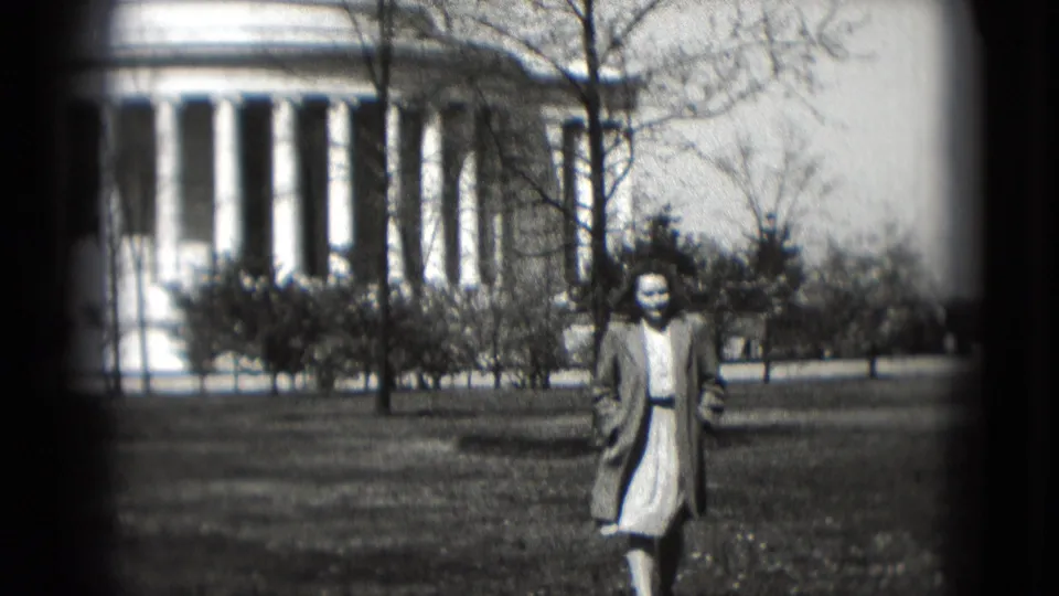 Black-and-white 8mm frame of a woman in a coat and dress on the lawn, with the Jefferson Memorial columns visible in the background.