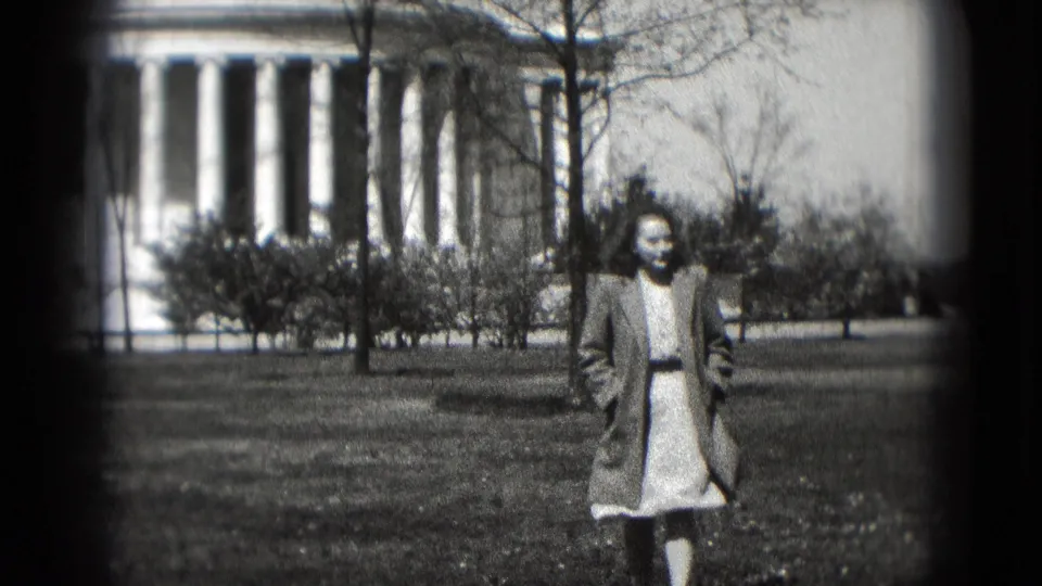 A woman in a long coat and dress stands near the camera on a Washington lawn, with the Jefferson Memorial behind her.