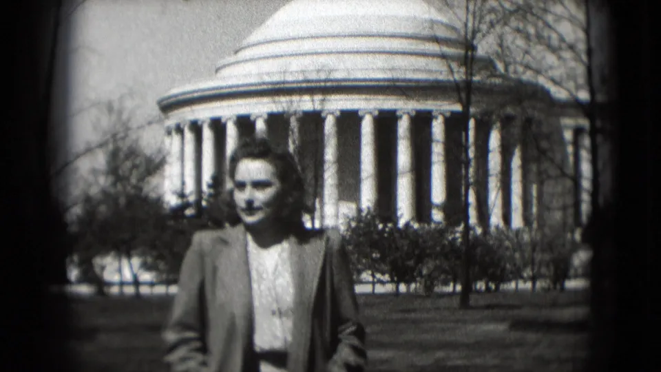 Adult woman walks toward the camera in front of the Jefferson Memorial, framed by lawn and bare trees.