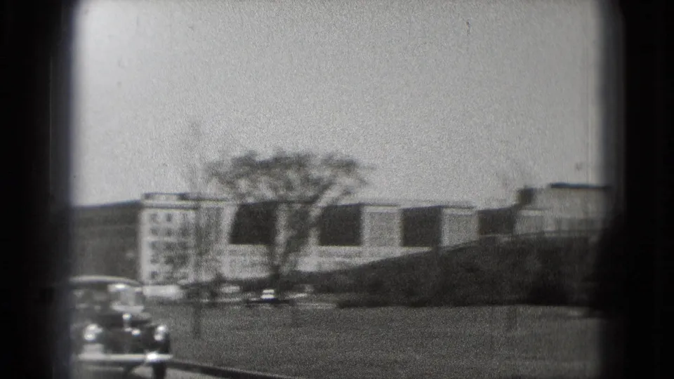 A Washington, DC government building appears across an open area, with older parked cars and a leafless tree in front.