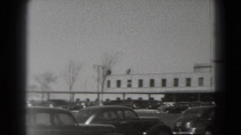 A broad federal building in Washington, DC is filmed past a row of parked 1970s-era cars and sparse trees.