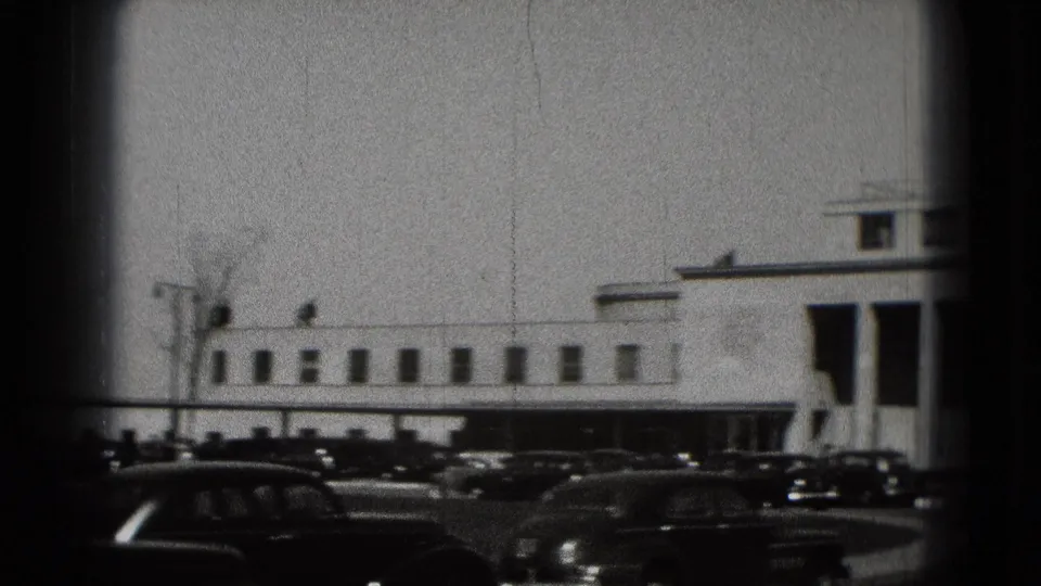 Washington, DC home-movie frame showing a long white modernist building and parked cars in the foreground.