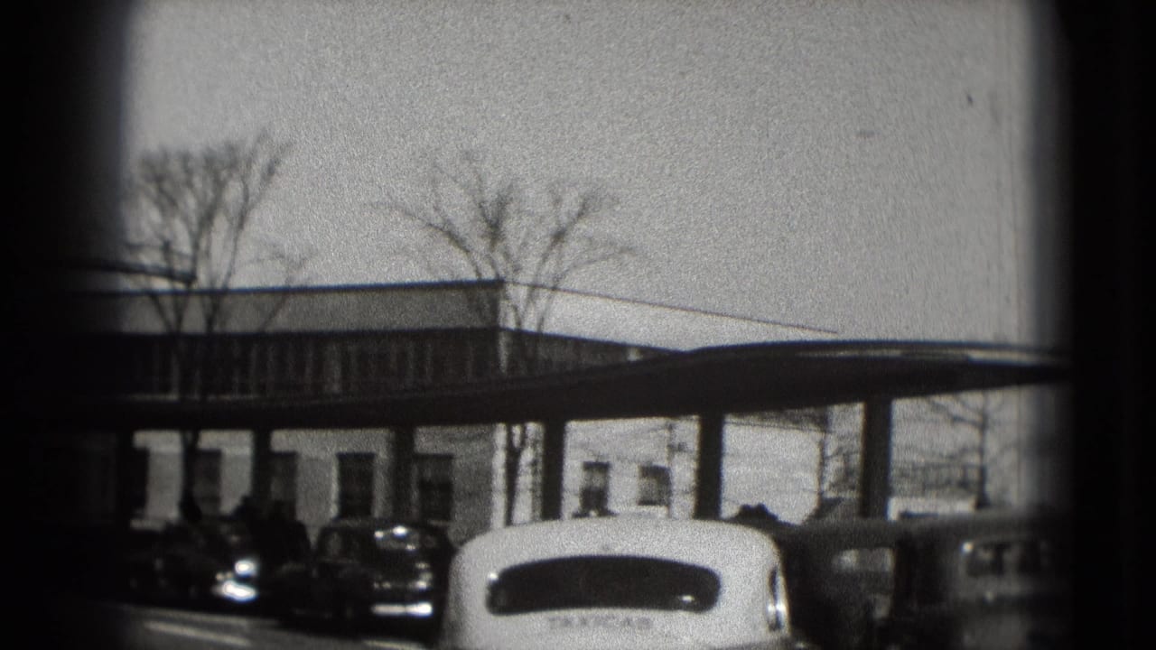 Washington DC home-movie frame from 1975 showing a low white building, leafless trees, and several parked mid-century cars.