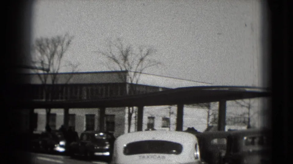 Washington DC home-movie frame from 1975 showing a low white building, leafless trees, and several parked mid-century cars.