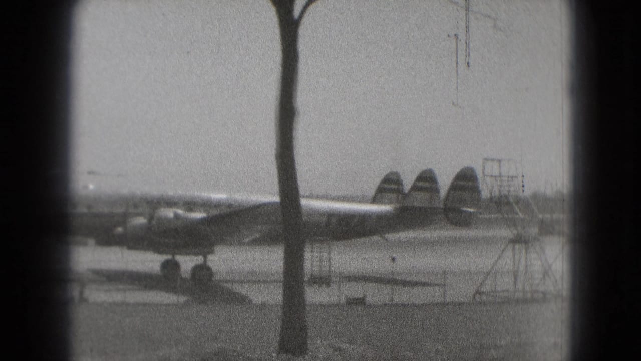 Propeller airplane on tarmac behind a tree, seen in grainy black-and-white film.