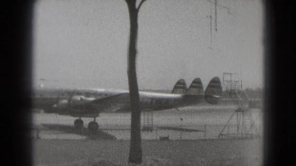 Washington DC 1975 home-movie frame showing a large propeller airplane partly blocked by a tree near open ground.