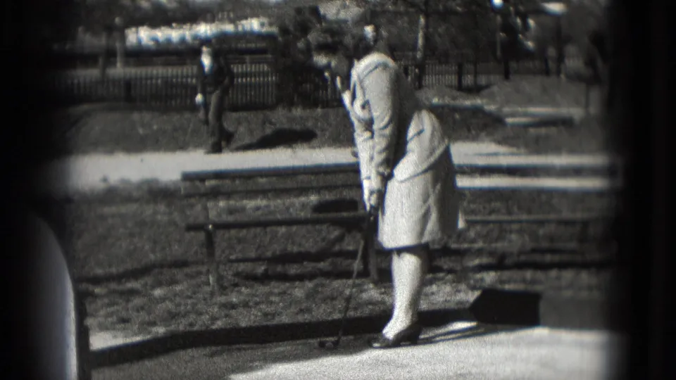 Washington DC 1975 frame of the woman completing a short putt near benches, with pedestrians and a fence in the background.