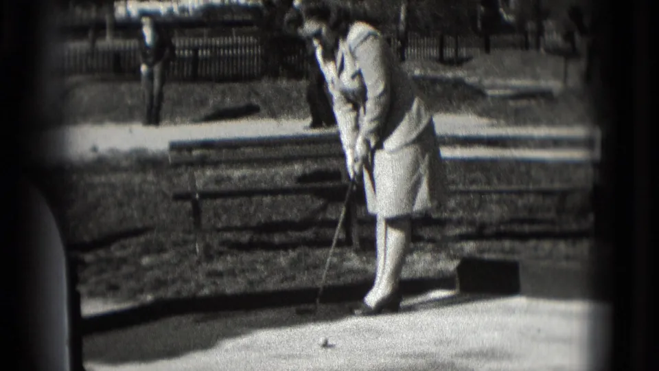 In supported 1975 Washington, DC home-movie footage, a woman in a light dress prepares a mini-golf putt on a sunny outdoor course.