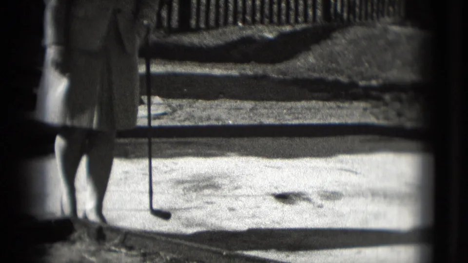 The camera frames the lower half of the woman with her putter and ball on the outdoor mini-golf lane in black-and-white 8mm footage.