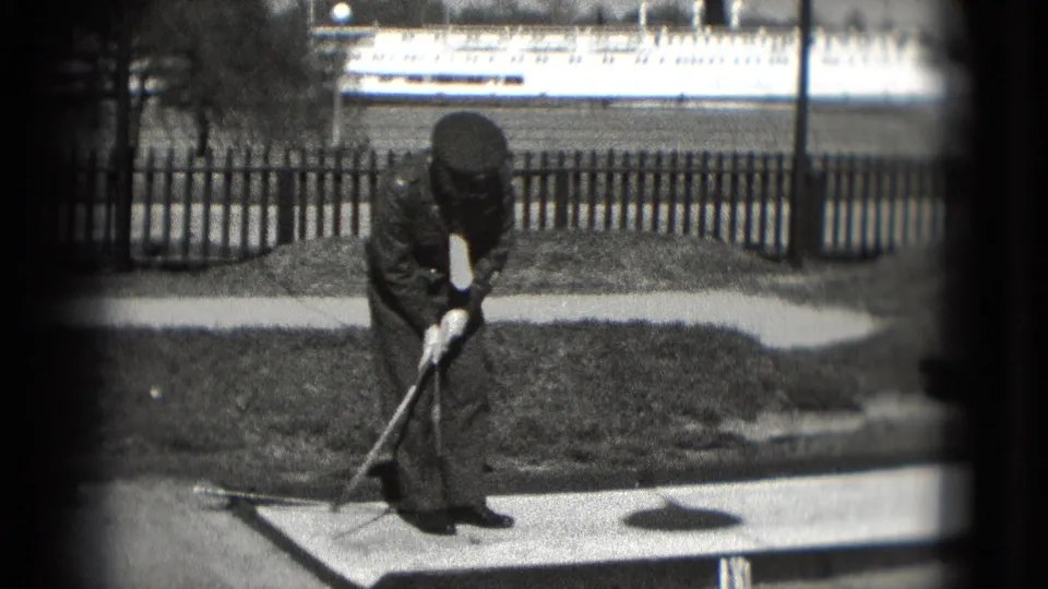 An adult in dark clothing, possibly in a uniform-style cap, bends to make a mini-golf shot near a fence in Washington, DC footage.