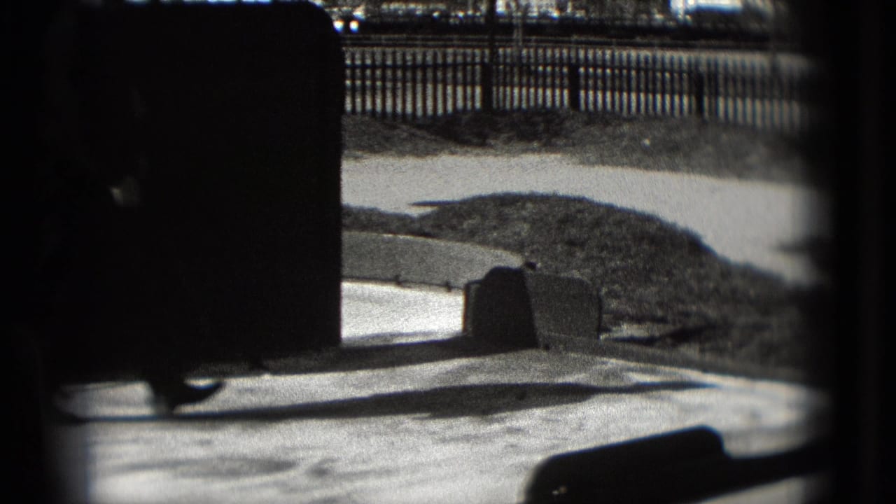 Paved mini-golf area seen through dark frame edges with fencing and low barriers in the background.