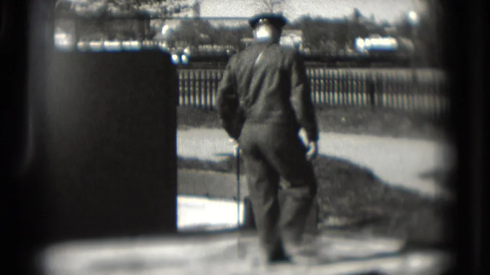 A uniformed man in 1975 Washington, DC walks across a small paved mini-golf course holding a club, seen through dark foreground edges.
