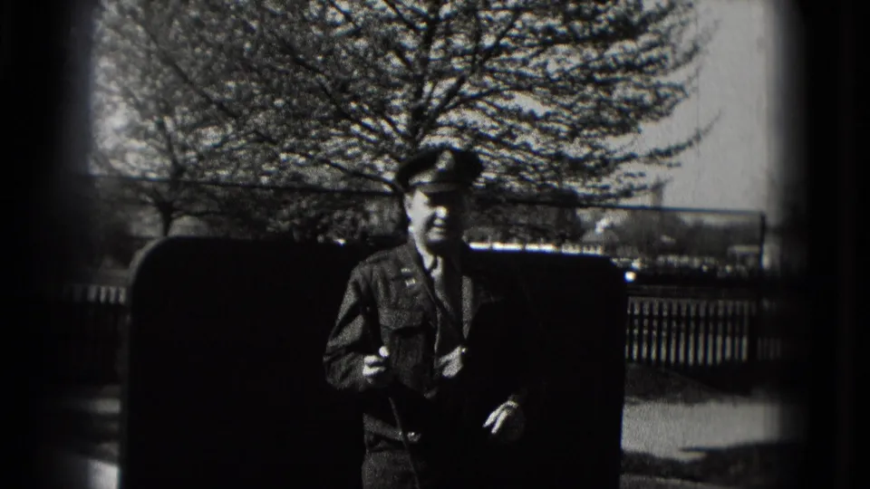 In 1975 Washington, DC home-movie footage, a suited man stands near an outdoor mini golf lane with fencing and trees behind him.