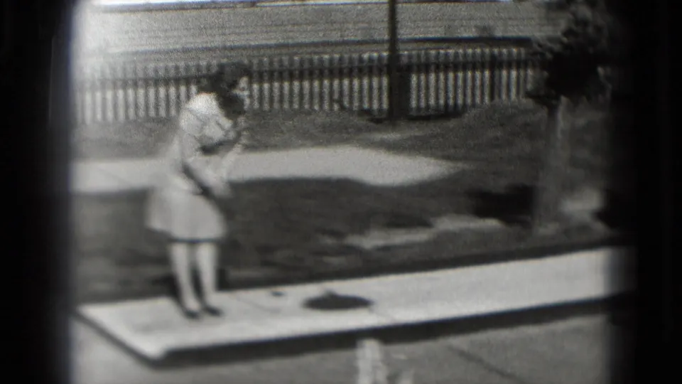 In 1975 Washington, DC footage, a woman in a light dress stands on a small outdoor mini golf lane near fencing and lawn.