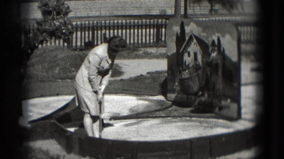 In 1975 Washington, DC home-movie footage, a woman bends to putt beside a painted house obstacle on an outdoor mini golf course.