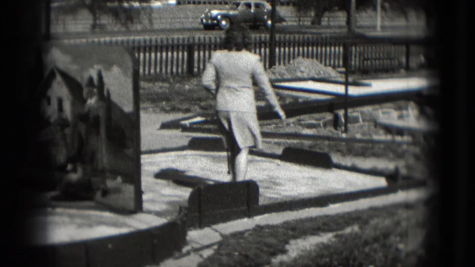 Adult woman walks between parts of the 1975 Washington DC chip-and-putt course, with a vintage car visible beyond the fence.