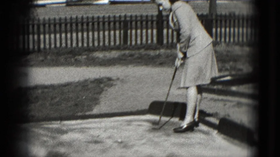 Adult woman bends over her club on a small chip-and-putt surface in 1975 Washington DC, with fence and grass in the background.