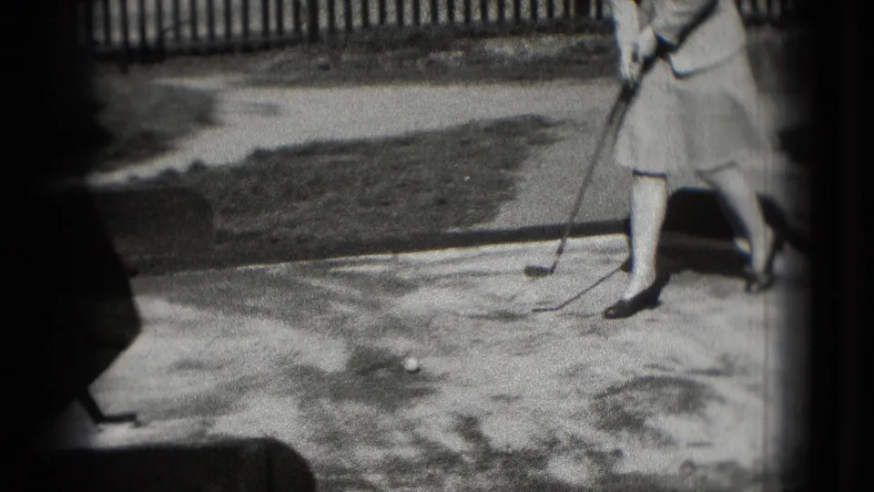 In Washington DC 1975 home-movie footage, a woman in a knee-length skirt and pumps prepares a putt near a wooden fence.