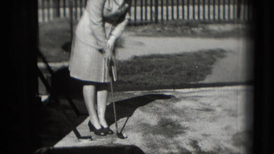 A woman in 1975 Washington DC footage bends over a putter on an outdoor putting surface, with her shadow crossing the ground.