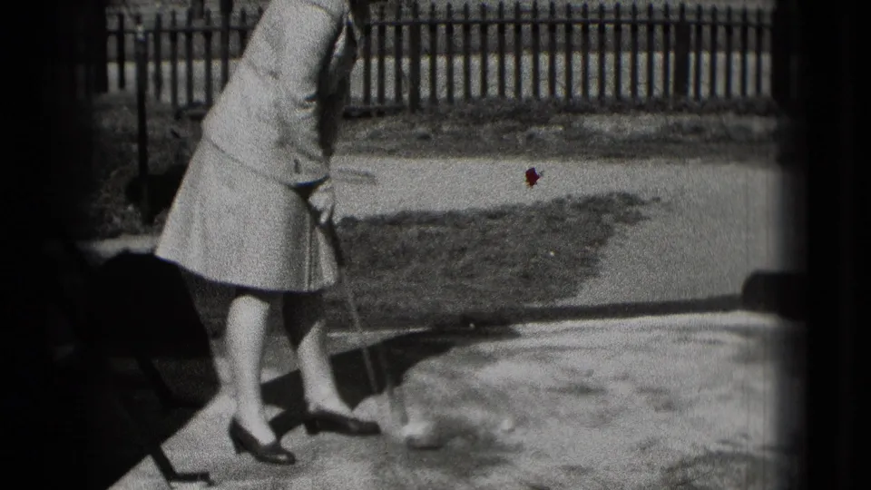 A 1975 Washington DC home-movie frame captures the woman after or during a putt, with the ball visible on the bright ground.