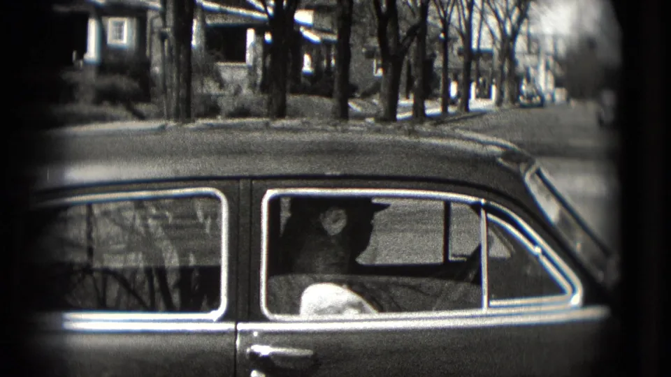 1975 Washington, DC frame with a hat-wearing driver seen through a sedan window on a residential street.