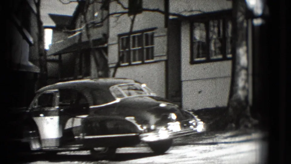 A rounded older sedan is parked along a Washington, DC residential curb beneath house windows in 1975 home-movie footage.