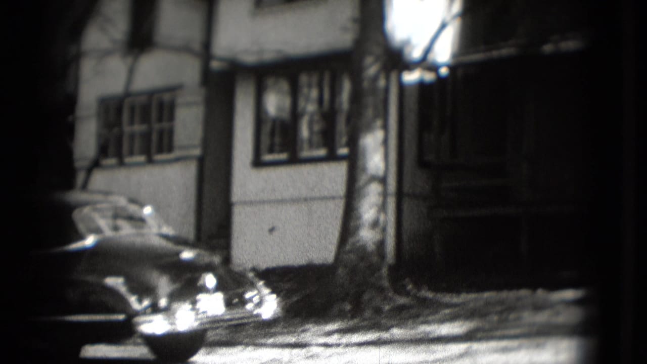 Parked vintage sedan beside a two-story house with bare trees and strong shadow.