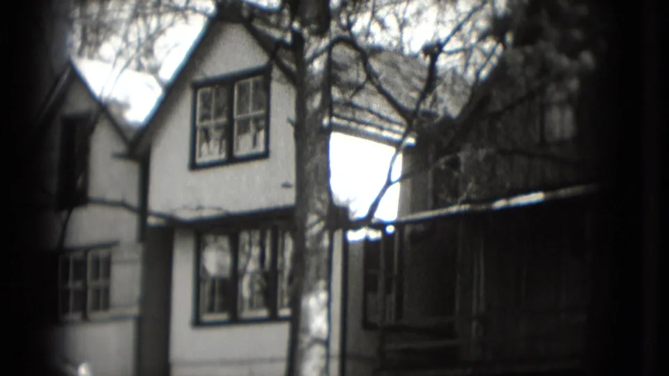 Black-and-white 8mm frame of a Washington DC residential house exterior with bare trees, windows, and dark trim or railing.