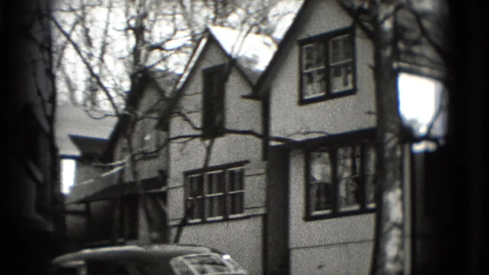 1975 Washington DC home-movie frame showing a mid-century sedan parked below a two-story house with bare branches overhead.
