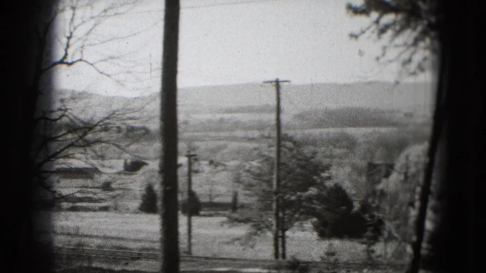 Black-and-white 8mm frame of open land near Washington, DC, with utility poles, bare branches, and low hills in the distance.