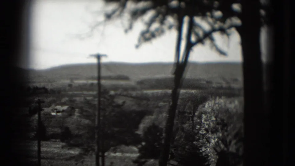 1975 8mm view across a soft rural hillside with utility poles, bare trees, and low buildings beneath a bright sky.