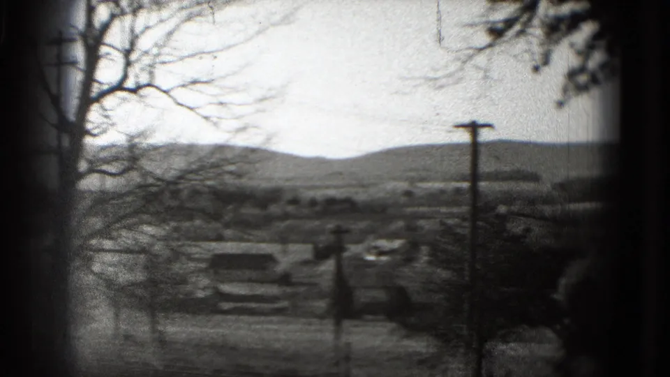 1975 8mm frame of open rural ground, a central utility pole, bare tree limbs, and hazy hills in the distance.