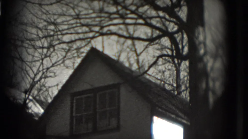 1975 8mm frame showing a pitched-roof house exterior behind bare tree branches, with dark frame edges and soft focus.