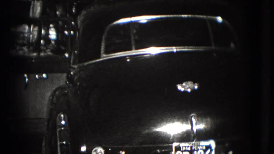 The camera shows more of the dark vintage sedan's rear body, chrome trim, and license plate in a shadowed outdoor setting.