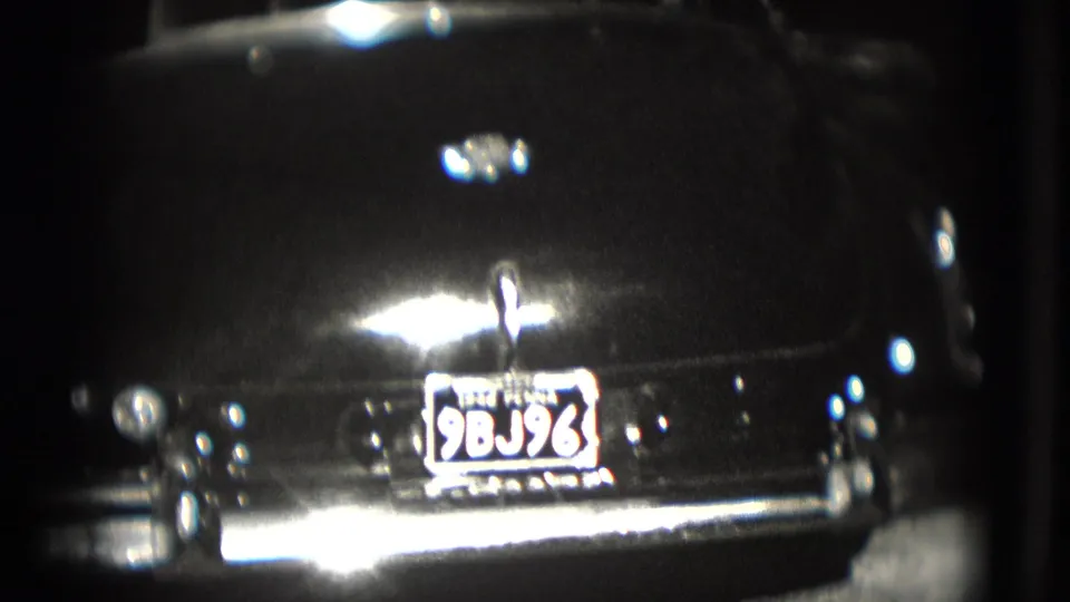 A closer rear view of the dark sedan shows the Pennsylvania plate, trunk emblem, bumper, and strong 8mm light flare.